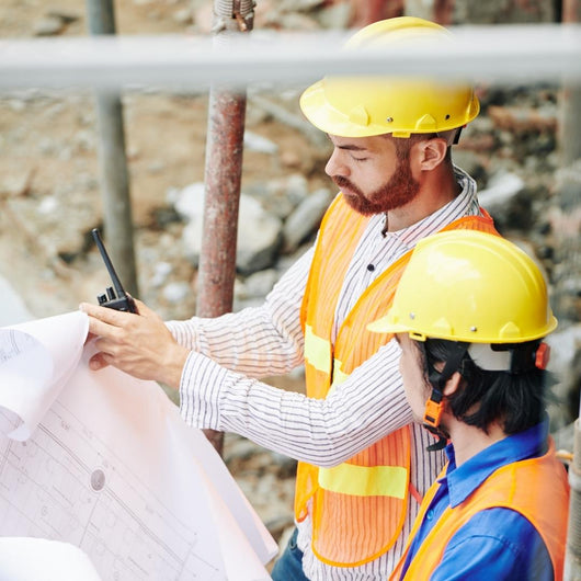 Two people wearing yellow hardhats, hold house plans.