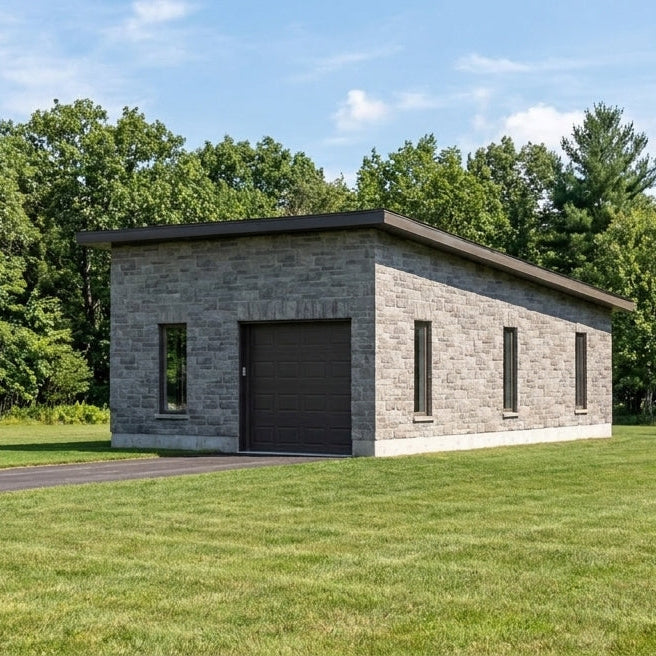 A modern single-car garage with a gray stone exterior and a dark garage door, situated on a green lawn with a paved driveway and a dense forest in the background under a blue sky.