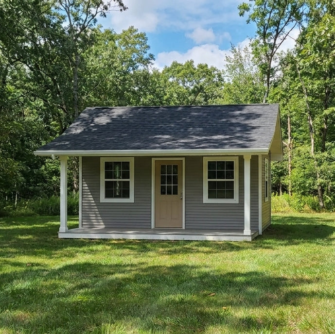 Exterior photo of House of Three Plan #21-0086, a one-storey she shed with grey horizontal siding, white trim, a half-glass door between two windows, a dark shingled gable roof and a full-width covered front porch with white columns.