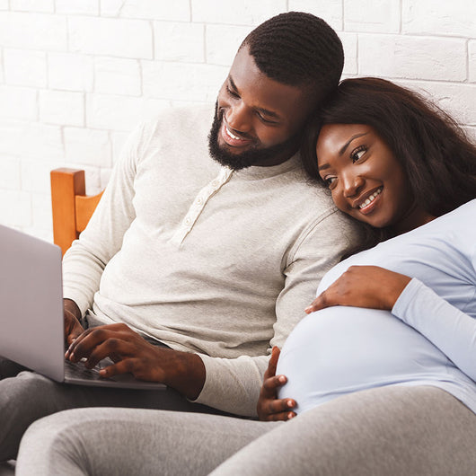 A man and woman sit together in front of a computer, smiling. The woman is visibly pregnant.